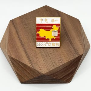 May include: A rectangular pin with a red and white background, featuring a yellow map of China and the text "5ESS®交换机". The pin also includes the AT&T logo and Chinese characters. The pin is set on a wooden, geometric-shaped base.