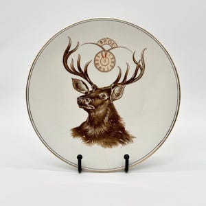 May include: A round ceramic plate featuring a brown illustration of a deer head with large antlers. Above the deer is a clock face with Roman numerals and the letters BPOE arched above it. The plate has a thin gold rim and a white background.