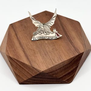 May include: A silver-colored pin featuring two dragons with outstretched wings, with the text "52nd COMBAT AVIATION". The pin is displayed on a geometric wooden block with a dark brown finish.