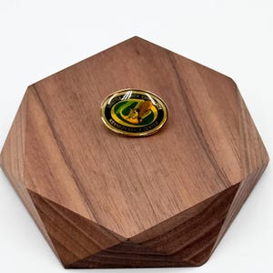 May include: An oval-shaped pin with a gold border, featuring a green and yellow design with the text "Shuttle Launch Experience" and "Kennedy Space Center." The pin is set on a geometric wooden display.