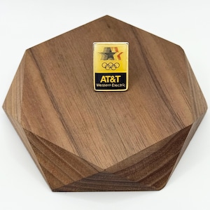 May include: A rectangular pin with a yellow background featuring the AT&T logo and Olympic rings. The pin has the text "AT&T Western Electric" in a dark blue rectangle. The pin is set on a wooden, geometric-shaped display.