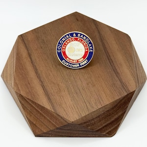 May include: A round pin with a blue, red, and white design. The pin has text that reads "COLONIAL & SANDLAKE, ORLANDO, FLORIDA" and "CUSTOMER FIRST". The pin is on a wooden, geometric-shaped stand.