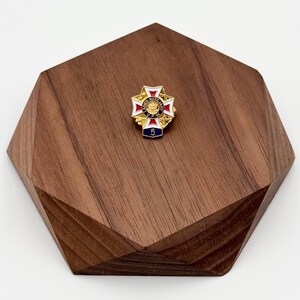 Puede incluir: Un pin dorado con un dise&ntilde;o de cruz, con detalles de esmalte rojo, blanco y azul. El pin tiene el texto "VFW AUXILIARY" y el n&uacute;mero "5" sobre un fondo azul. El pin se muestra sobre una base de madera geom&eacute;trica.