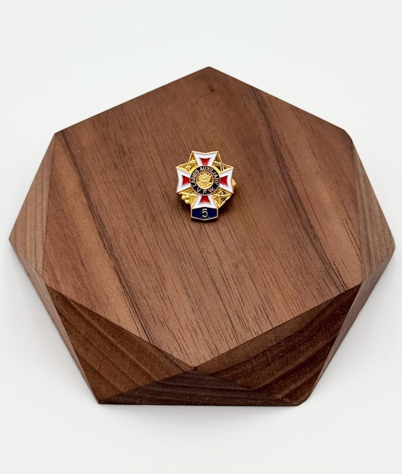 Puede incluir: Un pin dorado con un dise&ntilde;o de cruz, con detalles de esmalte rojo, blanco y azul. El pin tiene el texto "VFW AUXILIARY" y el n&uacute;mero "5" sobre un fondo azul. El pin se muestra sobre una base de madera geom&eacute;trica.