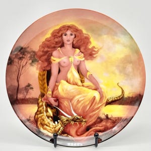 May include: A round decorative ceramic plate featuring a detailed illustration of a person with long red hair wearing a yellow garment, seated beside a golden dragon. The background depicts a sunset landscape with trees and a body of water.