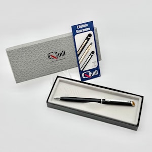 May include: A black ballpoint pen with silver accents rests in a rectangular black box with a light gray interior. A textured gray box lid and a blue informational card featuring the text Lifetime Guarantee and Quill are positioned behind the pen.