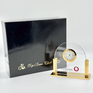May include: A quartz clock with a gold-toned circular face and Roman numerals, mounted on a clear arched acrylic panel with gold-toned fluted pillars and a rectangular base. The clock is displayed next to a black box labeled Top Brass Clock.