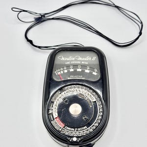 May include: A black Weston Master II cine exposure meter featuring a curved scale for relative brightness and a circular dial for camera f-setting adjustments. The device includes a thin black neck strap attached to the top of the casing.