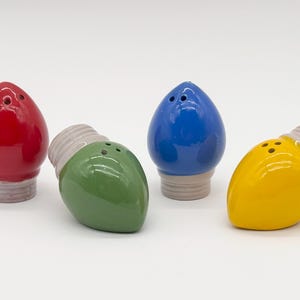 May include: Set of four ceramic salt and pepper shakers shaped like Christmas light bulbs. The bulbs are red, green, blue, and yellow, with gray bases and bulb sockets. Each shaker has three holes for dispensing.