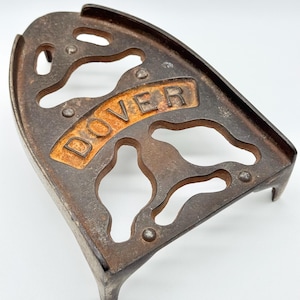 May include: A vintage cast iron trivet with a dark, weathered finish and an openwork design. The word DOVER is embossed in raised lettering across the center. The object features a pointed, triangular shape with three small feet for support.