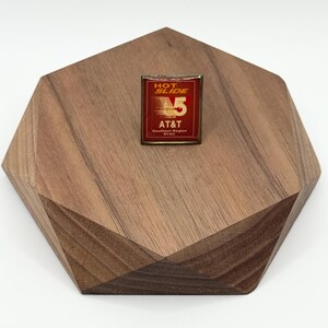May include: A vintage pin with the text "HOT SLIDE 5" and "AT&T Southern Region RTAC" on a red background. The pin is set on a geometric wooden display piece with a light brown finish.