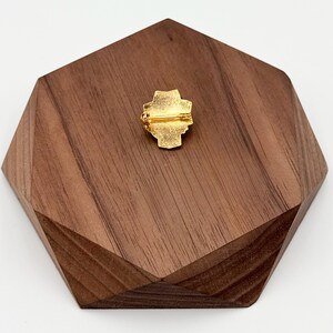 Puede incluir: Un broche dorado con un dise&ntilde;o en forma de cruz, colocado sobre una base geom&eacute;trica de madera oscura. La parte trasera del broche tiene un cierre para una fijaci&oacute;n segura. La base de madera tiene una forma &uacute;nica y multifac&eacute;tica.