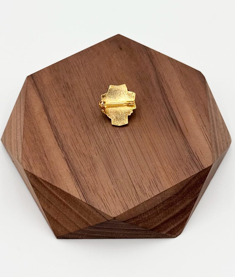 Puede incluir: Un broche dorado con un dise&ntilde;o en forma de cruz, colocado sobre una base geom&eacute;trica de madera oscura. La parte trasera del broche tiene un cierre para una fijaci&oacute;n segura. La base de madera tiene una forma &uacute;nica y multifac&eacute;tica.