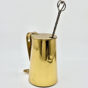 May include: A polished brass wall-mounted container with a hinged lid and a vertical handle featuring a keyhole slot for mounting. A long, twisted metal rod with a looped top extends through a small circular opening in the lid of the vessel.