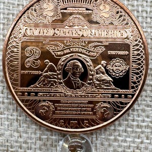 May include: A copper-colored United States Two Silver Dollars coin. The coin features an image of George Washington, the text "United States of America", and other intricate designs. A clear, round object is below the coin.