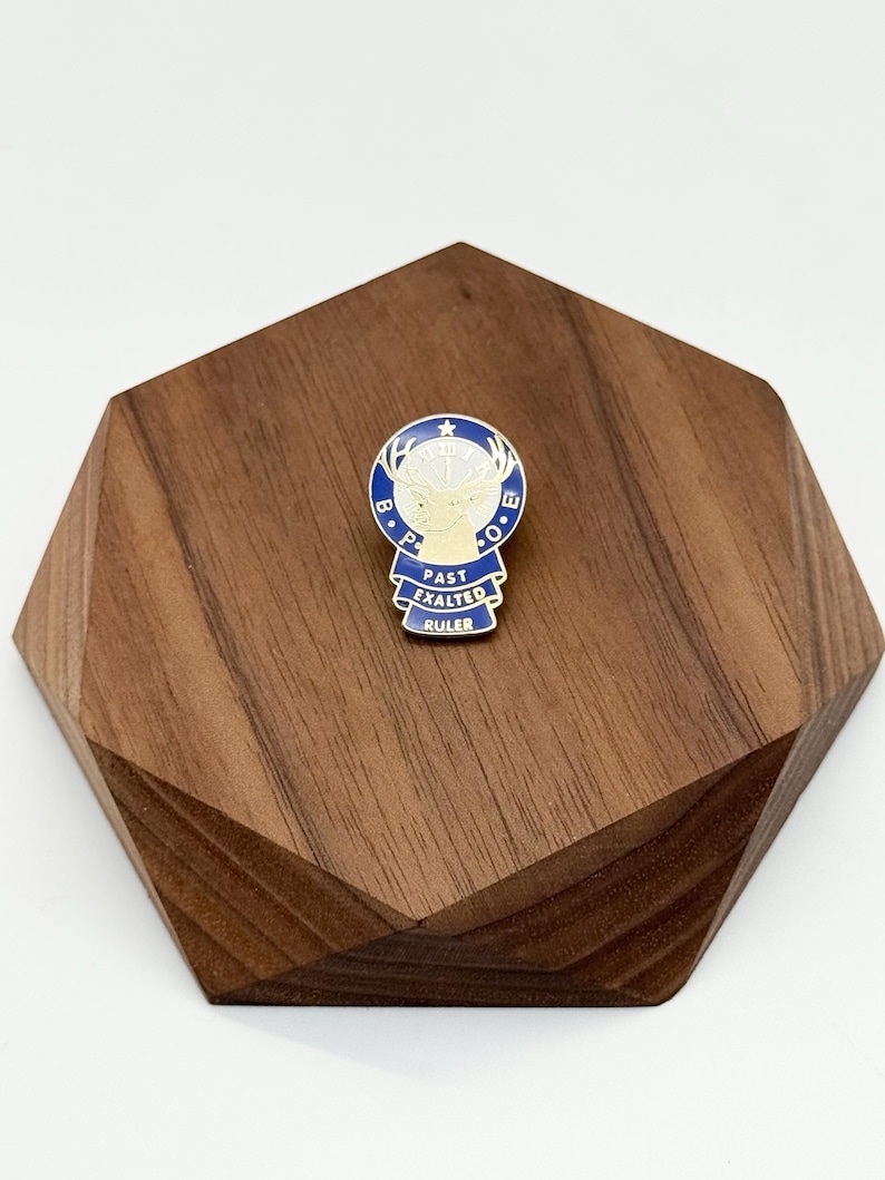 Puede incluir: Un pin azul, blanco y dorado con el texto "PAST EXALTED RULER" y un emblema de cabeza de ciervo. El pin se muestra sobre un soporte de madera geom&eacute;trico marr&oacute;n oscuro.