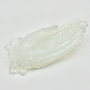 May include: A translucent, milky white glass sculpture shaped like an open hand with fingers extended. The wrist area features a molded floral detail with petals. The object has a smooth, glossy finish and a slightly iridescent, opalescent quality.