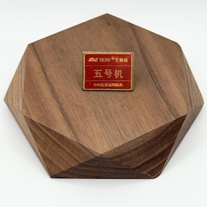 May include: A small, rectangular pin with red and gold text, reading "AT&T 5ESS" and Chinese characters, sits on a geometric wooden display. The pin is a vintage collectible, likely a promotional item or souvenir.