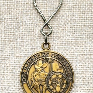 May include: A vintage gold-toned keychain featuring a round medallion with the text "1992 B.P.O. Elks Convention Dallas, Texas." The medallion depicts a cowboy figure and a deer head. The keychain has a decorative loop and a clear crystal at the top.