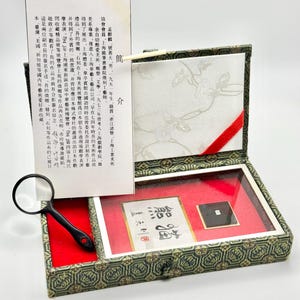 May include: A rectangular box with a patterned green fabric exterior, containing a red velvet-lined interior with a small square black object, a magnifying glass, and a white card featuring Chinese text and a small illustration of a bird.