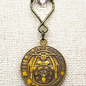 May include: A vintage gold-toned pendant with a decorative loop and a small, clear bead. The pendant features the text "1991 B.P.O. Elks Convention St. Louis, Missouri" surrounding a central emblem of a deer head and other symbols.