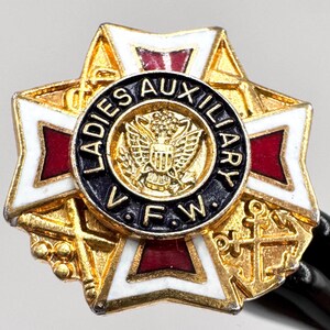 May include: A gold-toned Ladies Auxiliary VFW pin. The pin features a starburst design with white, red, and gold accents. The center has a black circle with the words "Ladies Auxiliary VFW" and an emblem.