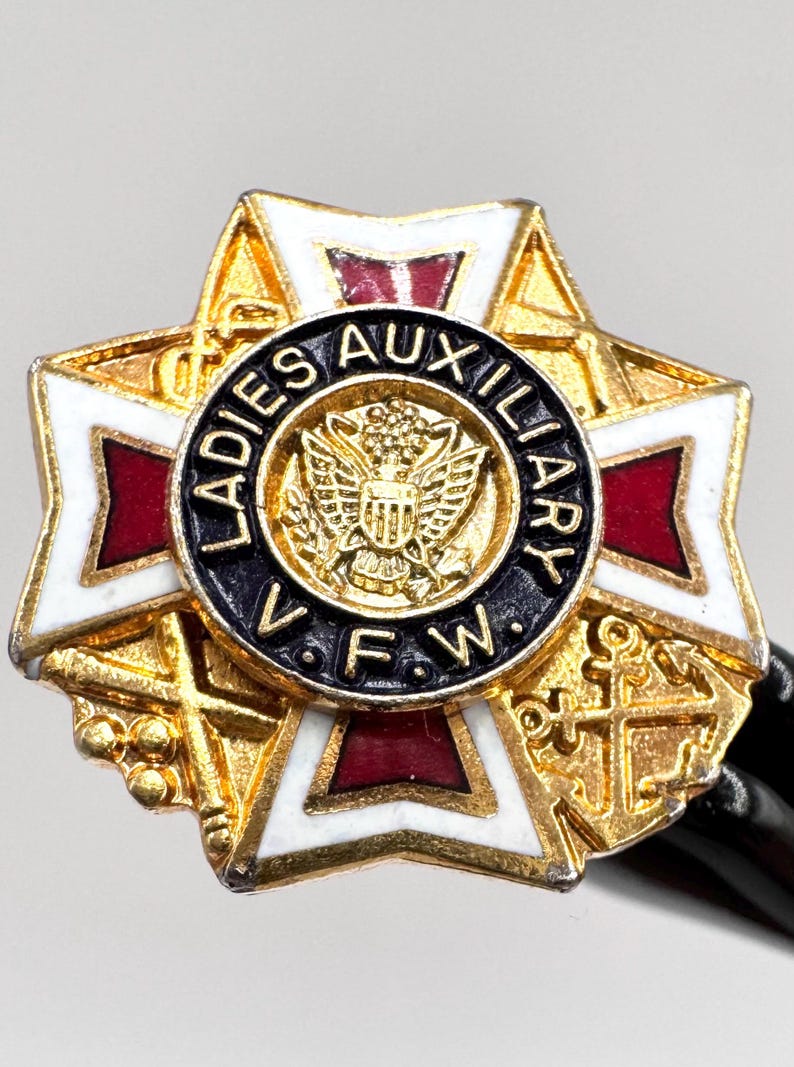 May include: A gold-toned Ladies Auxiliary VFW pin. The pin features a starburst design with white, red, and gold accents. The center has a black circle with the words "Ladies Auxiliary VFW" and an emblem.
