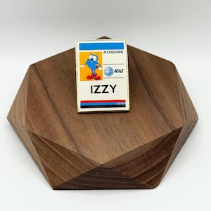 May include: A rectangular pin featuring the AT&T Izzy character. The pin has a blue and white top section with the text "AO80496". Below is a graphic of Izzy, the AT&T logo, and the name "IZZY" in bold letters. The pin is set on a wooden geometric display.