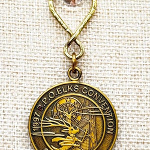 May include: A vintage gold-toned pendant with a loop and a small clear bead. The pendant features the text "1997 B.P.O. ELKS CONVENTION Chicago, Illinois" around a detailed image of a deer and a clock tower.