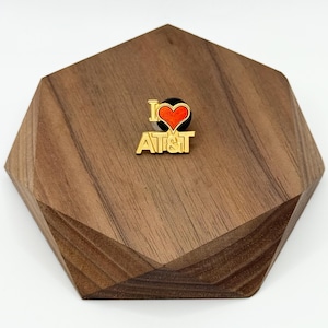 May include: A gold-colored pin with the text "I ♥ AT&T" and a red heart. The pin is set on a dark brown, geometric wooden display piece. The background is white.