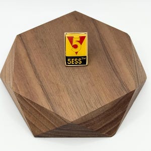 May include: A rectangular enamel pin with a gold border, featuring a red number 5 inside a yellow triangle, set against a black background. The text "5ESS™" is below the number. The pin is displayed on a wooden, geometric-shaped coaster.