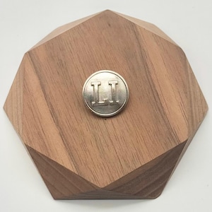 May include: A circular silver-toned metal button featuring the embossed capital letters LI in a serif font. The button is centered on a faceted, geometric wooden block with a natural brown wood grain finish and visible angled edges.