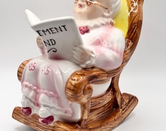May include: A ceramic figurine of a woman in a rocking chair, reading a book. The figurine is painted with a glossy finish, featuring a white head, pink dress, and brown rocking chair. The book reads "EMENT ND".