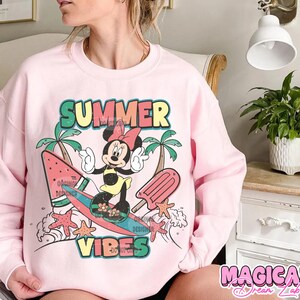 May include: Pink sweatshirt with a graphic of Mickey Mouse surfing on a watermelon surfboard. The graphic also includes palm trees, a popsicle, and the text "Summer Vibes".