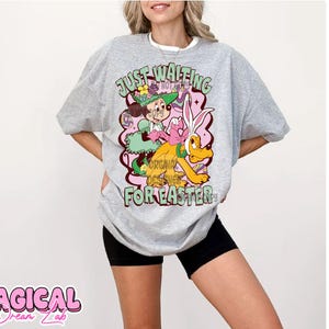 Puede incluir: Una camiseta gris con un estampado gr&aacute;fico de un conejo de dibujos animados y una ni&ntilde;a que sostiene una cesta de huevos. El texto "Just Waiting For Easter" est&aacute; impreso en la camiseta.