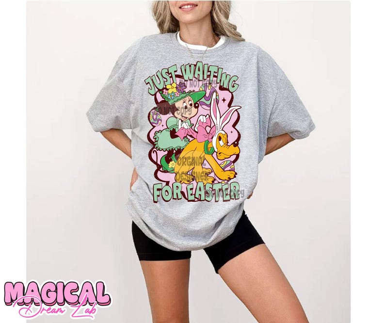 Puede incluir: Una camiseta gris con un estampado gr&aacute;fico de un conejo de dibujos animados y una ni&ntilde;a que sostiene una cesta de huevos. El texto "Just Waiting For Easter" est&aacute; impreso en la camiseta.