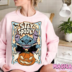 May include: A light pink sweatshirt featuring a Halloween-themed graphic. The design includes the text "STAY SPOOKY" above a cartoon character, a jack-o'-lantern, and two ghosts. The sweatshirt is a comfortable, casual style.