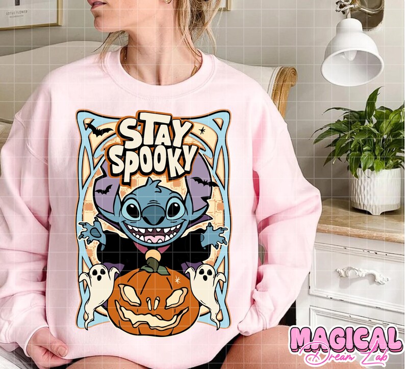 May include: A light pink sweatshirt featuring a Halloween-themed graphic. The design includes the text "STAY SPOOKY" above a cartoon character, a jack-o'-lantern, and two ghosts. The sweatshirt is a comfortable, casual style.