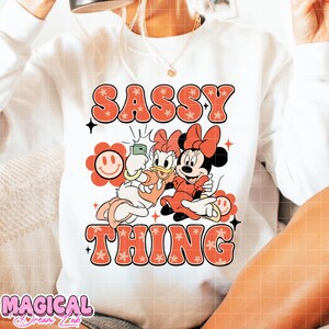 Puede incluir: Sudadera blanca con las palabras "SASSY THING" en letras burbuja naranjas con detalles florales. El dise&ntilde;o presenta a Daisy Duck y Minnie Mouse haci&eacute;ndose un selfie. Tambi&eacute;n incluye un gr&aacute;fico de una flor sonriente.