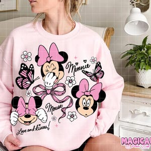 Peut inclure: Un sweat-shirt rose clair avec des graphiques de Minnie Mouse. Le design comprend le visage de Minnie, des n&oelig;uds, des papillons, des fleurs et les mots "Minnie", "Mouse" et "Love and kisses!".