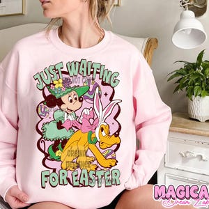 Puede incluir: Sudadera rosa con un gr&aacute;fico de Mickey Mouse y Pluto en un dise&ntilde;o de Pascua. El texto "Just Waiting for Easter" est&aacute; impreso en la sudadera.
