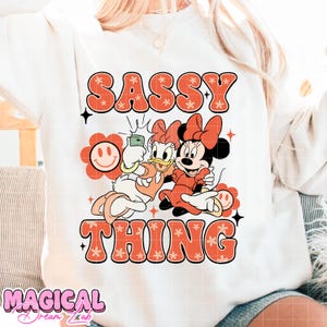 Puede incluir: Sudadera blanca con las palabras "SASSY THING" en naranja, con letras de estilo floral. El gr&aacute;fico presenta a Daisy Duck y Minnie Mouse haci&eacute;ndose un selfie. Tambi&eacute;n se ve una flor sonriente y las palabras "MAGICAL Dreams Lab".