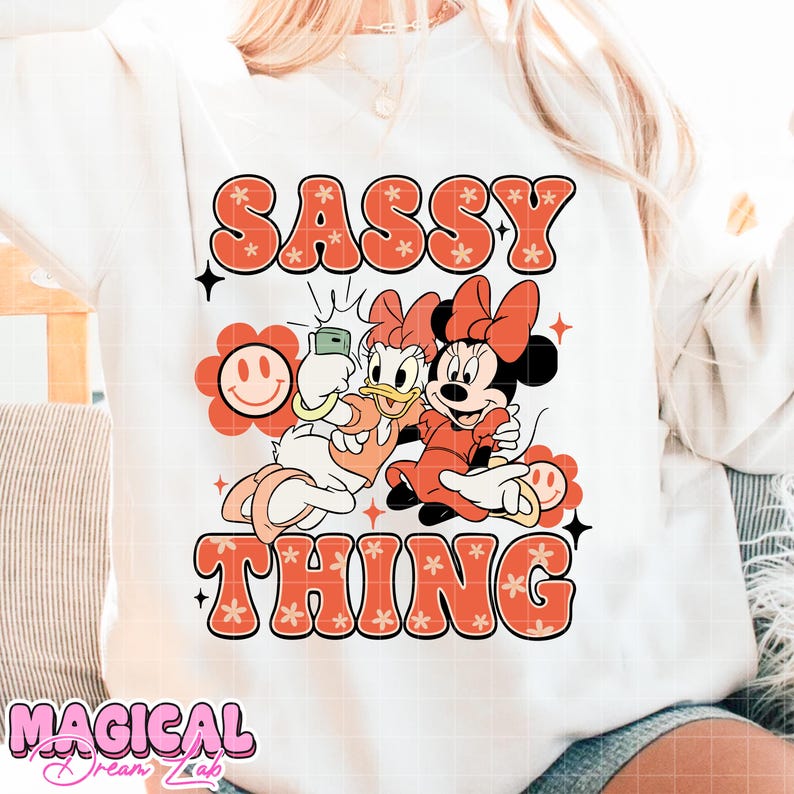 Puede incluir: Sudadera blanca con las palabras "SASSY THING" en naranja, con letras de estilo floral. El gr&aacute;fico presenta a Daisy Duck y Minnie Mouse haci&eacute;ndose un selfie. Tambi&eacute;n se ve una flor sonriente y las palabras "MAGICAL Dreams Lab".