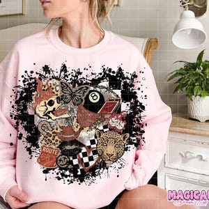 May include: Light pink sweatshirt with a graphic design featuring a skull, butterfly, leopard, Route 66 sign, and an 8-ball. The design is surrounded by black paint splatters. The text "MAGICAL Dream Lab" is in the bottom right corner.