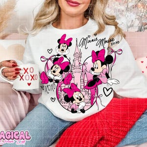May include: White long-sleeved sweatshirt featuring a Minnie Mouse design in pink and black. The design includes Minnie Mouse characters, hearts, and the text "Minnie Mouse XOXO". A white mug with "XO XOXO" is held in the hand.