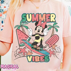 May include: Pink t-shirt with a graphic of Minnie Mouse surfing on a watermelon surfboard. The text "SUMMER VIBES" is printed in colorful letters on the shirt.