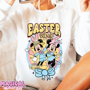 May include: White sweatshirt with a colorful Easter design featuring Mickey and Minnie Mouse in pastel colors. The text "Easter Vibes" is written in a retro font.