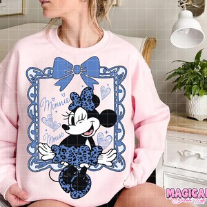 May include: A light pink sweatshirt featuring a Minnie Mouse graphic. The graphic includes a blue bow, a framed image of Minnie Mouse, and the words "Minnie Mouse". The Minnie Mouse image has a blue leopard print bow and skirt.