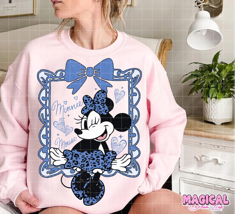 May include: A light pink sweatshirt featuring a Minnie Mouse graphic. The graphic includes a blue bow, a framed image of Minnie Mouse, and the words "Minnie Mouse". The Minnie Mouse image has a blue leopard print bow and skirt.