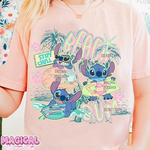 May include: A light pink t-shirt featuring a colorful graphic of the character Stitch at the beach. The design includes the words "BEACH SUN" and "STAY CHILL". The shirt has short sleeves and a crew neck.